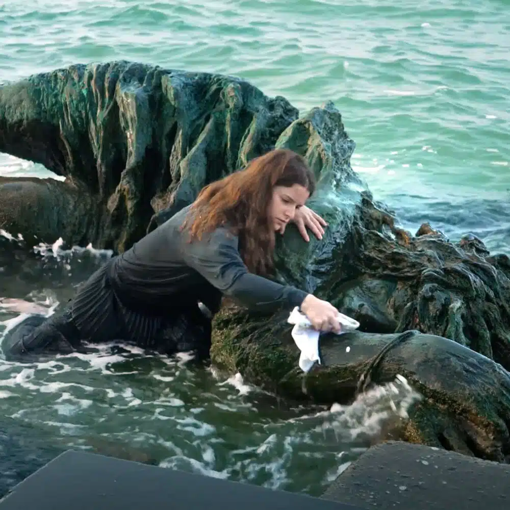 Detail of a work by Edith Dekyndt depicting a woman in the water with a cloth cleaning a monumental statue lying in the waves. 